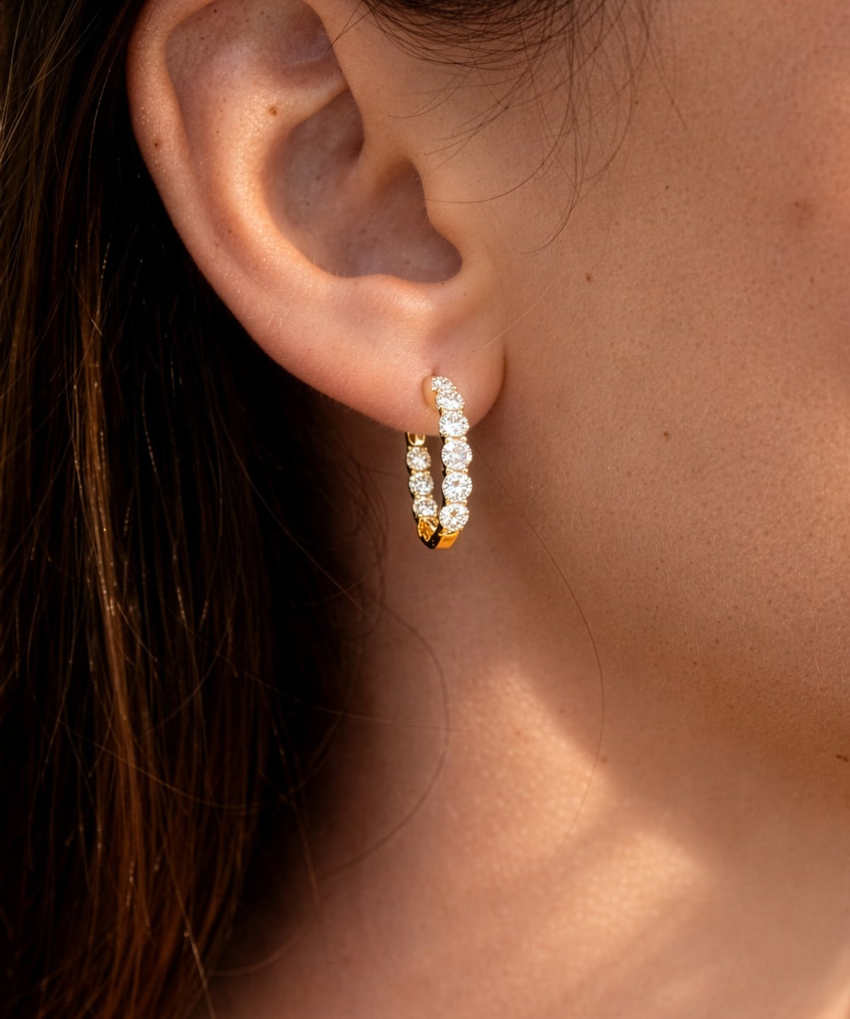 Lady's ear wearing a pair of yellow gold hoop earrings with clear lab grown diamonds on a white background
