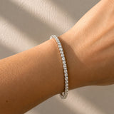 A white tennis bracelet with clear lab diamonds worn on a person's wrist, shown against a neutral beige background with soft diagonal shadows.