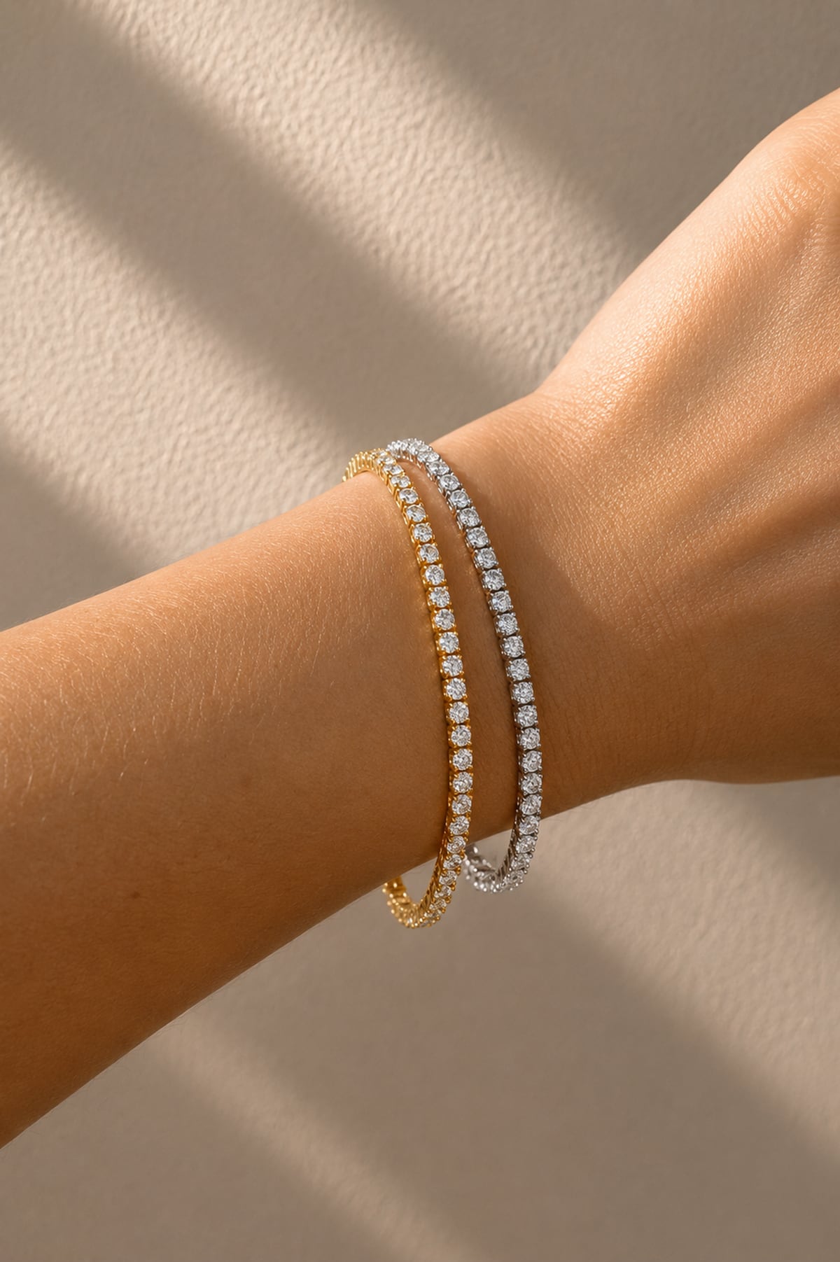 Two slim tennis bracelets on a person's wrist, one in yellow gold and one in silver, against a softly lit neutral background.