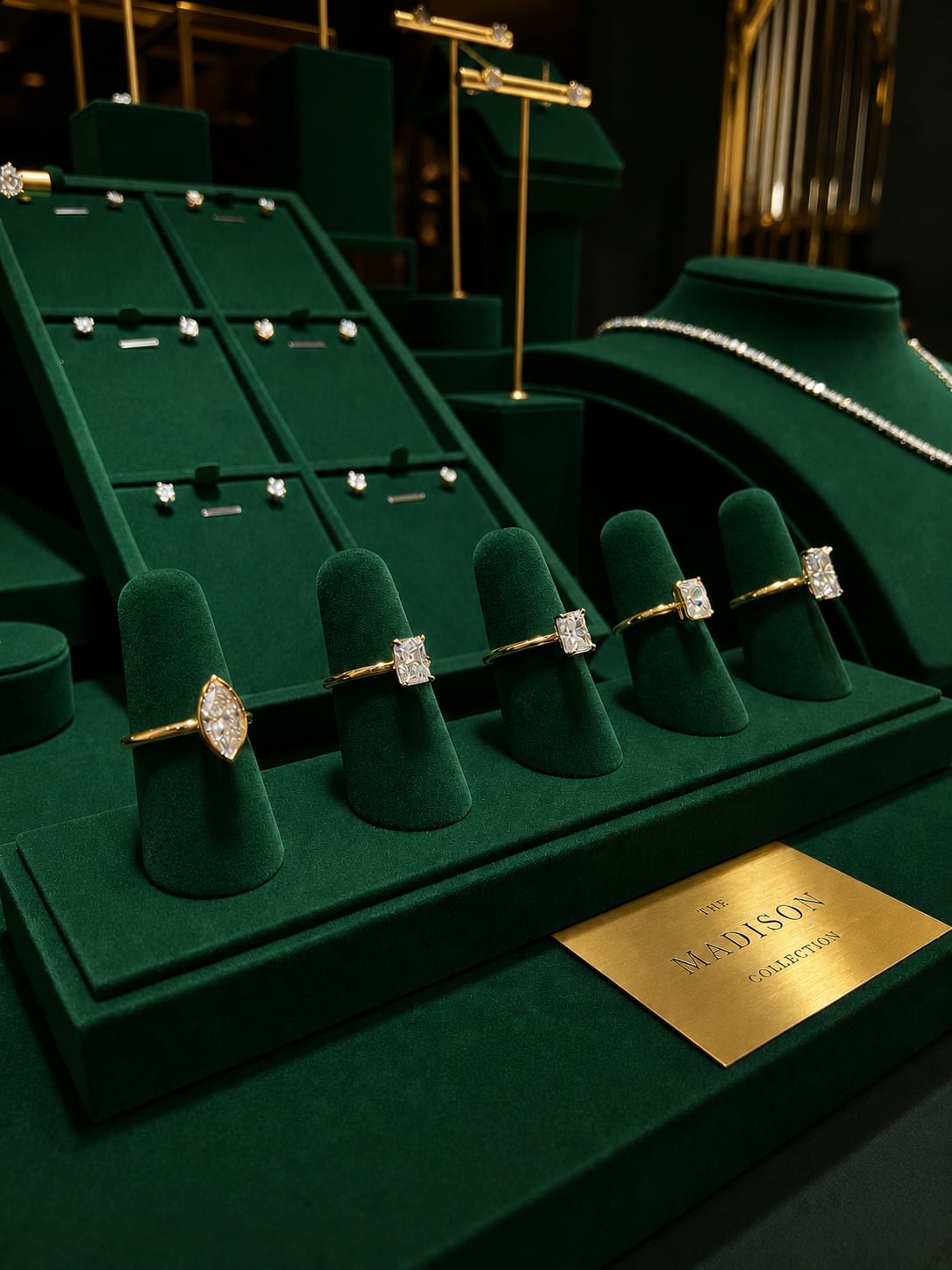 A luxury jewelry display featuring yellow gold rings with diamond settings on dark green velvet display stands, alongside a matching diamond necklace on a green display bust. A gold brand plaque for 'The Madison Collection' is visible in the foreground.