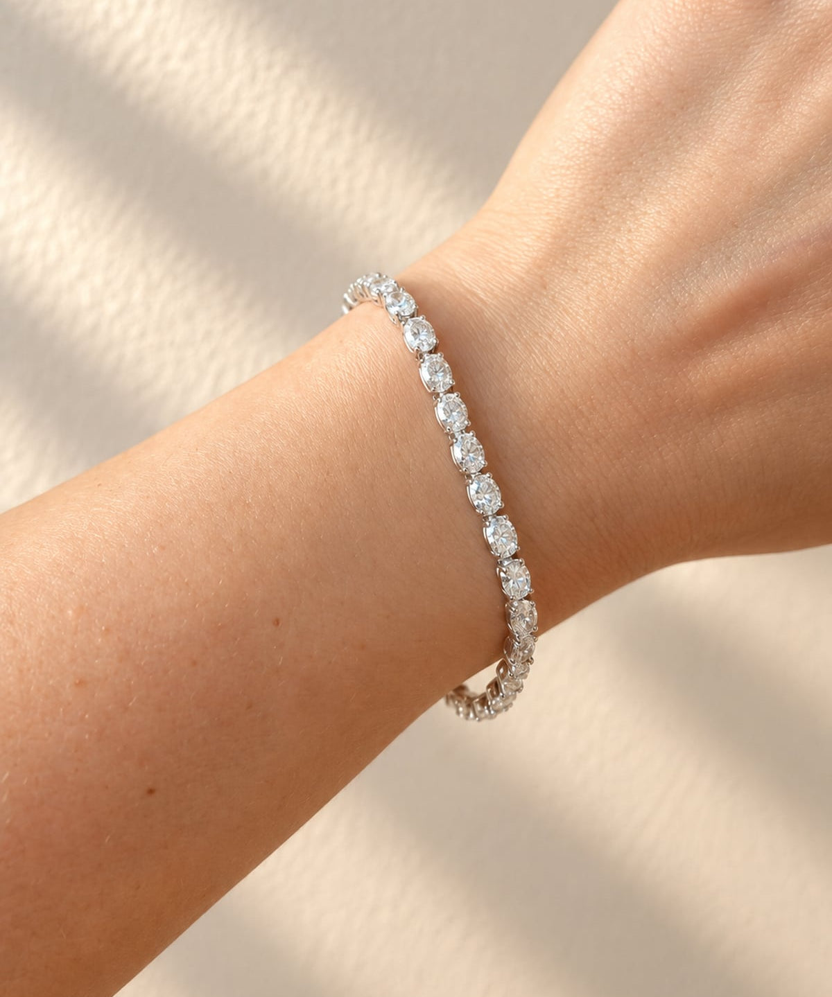 A close-up of a person's wrist wearing a delicate silver platinum bracelet with sparkling clear diamonds, set against a warm beige background with soft shadow patterns.